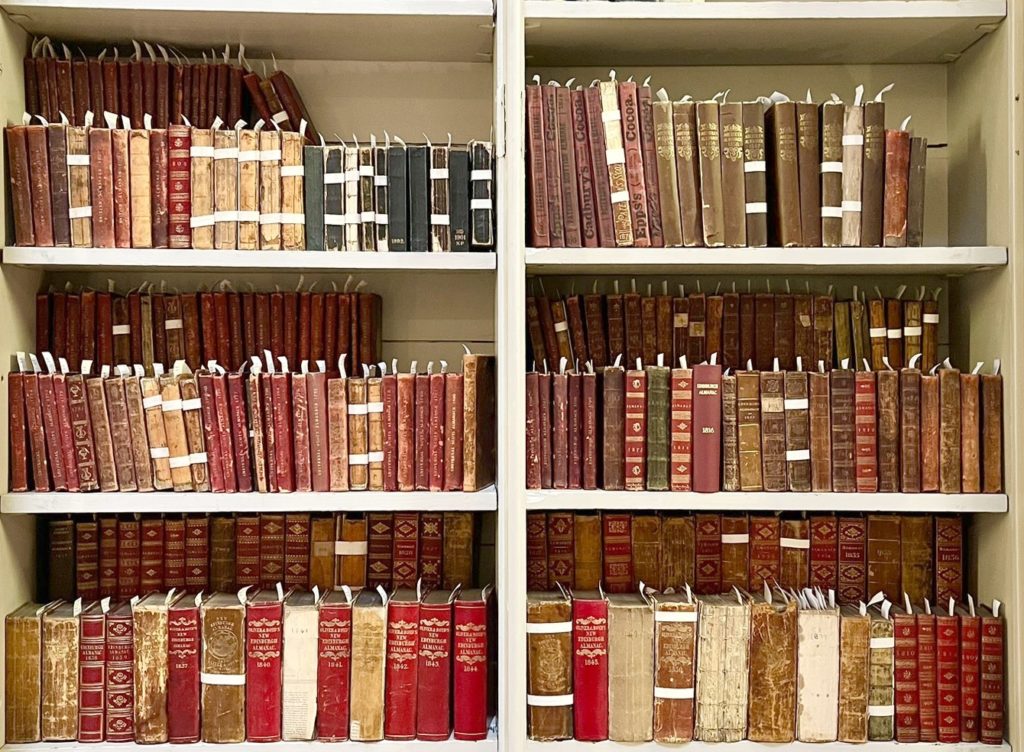 Almanacs bound in red leather and black cloth on white shelves.